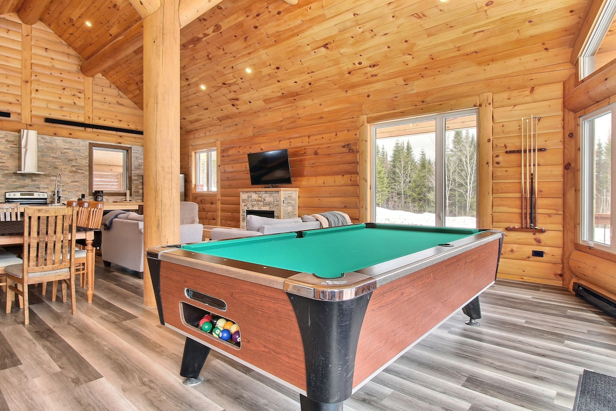 A spacious game room is highlighted by a billiard table surrounded by wooden beams and warm-toned walls. Large windows provide natural light and views of the outdoor scenery. A dining table and comfortable seating area can be seen in the background, enhancing the inviting space.