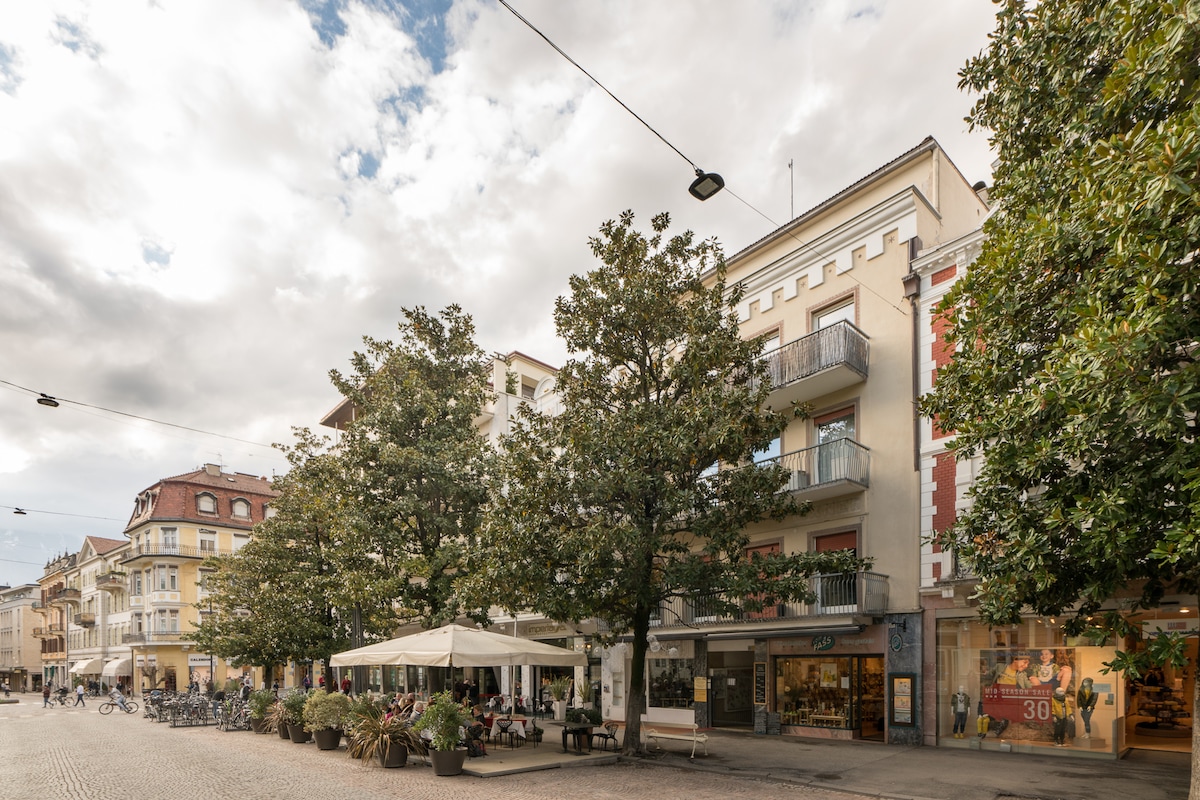 A lively pedestrian zone is depicted, featuring several buildings adorned with balconies. Trees provide greenery, while outdoor seating areas are visible, suggesting a welcoming atmosphere for visitors. The scene captures a blend of urban life with a tranquil ambiance under a partly cloudy sky.