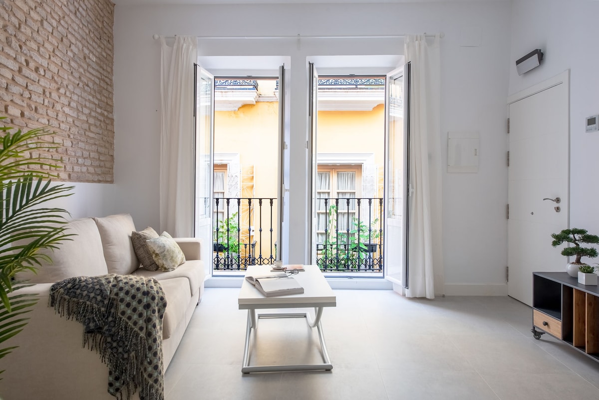 A light-filled living area features a soft sofa adorned with decorative pillows. Two large windows open to a view of a charming yellow building, allowing natural light to enhance the space. A minimalist coffee table occupies the center, complemented by a simple plant nearby.