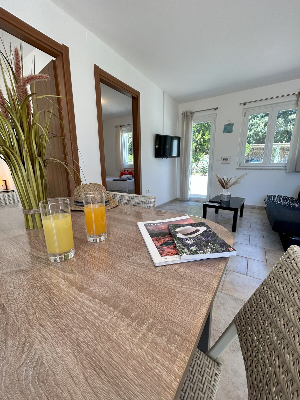 A dining area is presented with two glasses of juice, complemented by a magazine placed on the table. In the background, a cozy living space with a television and large windows offers views to the outside, allowing natural light to enhance the room.