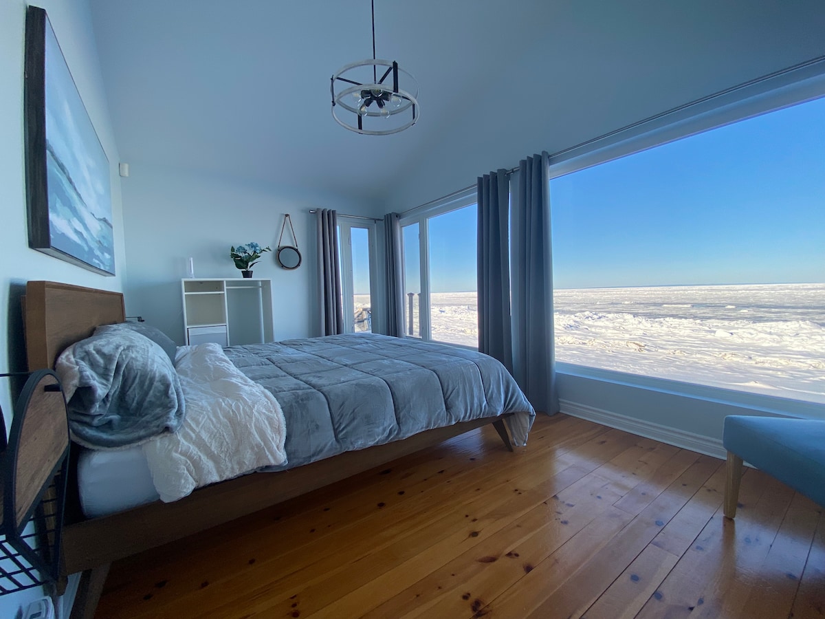 A bright bedroom features large windows that provide an ocean view, flooding the space with natural light. A spacious bed with layered bedding is positioned against the wall. Wooden flooring and a stylish light fixture complement the minimalist décor.