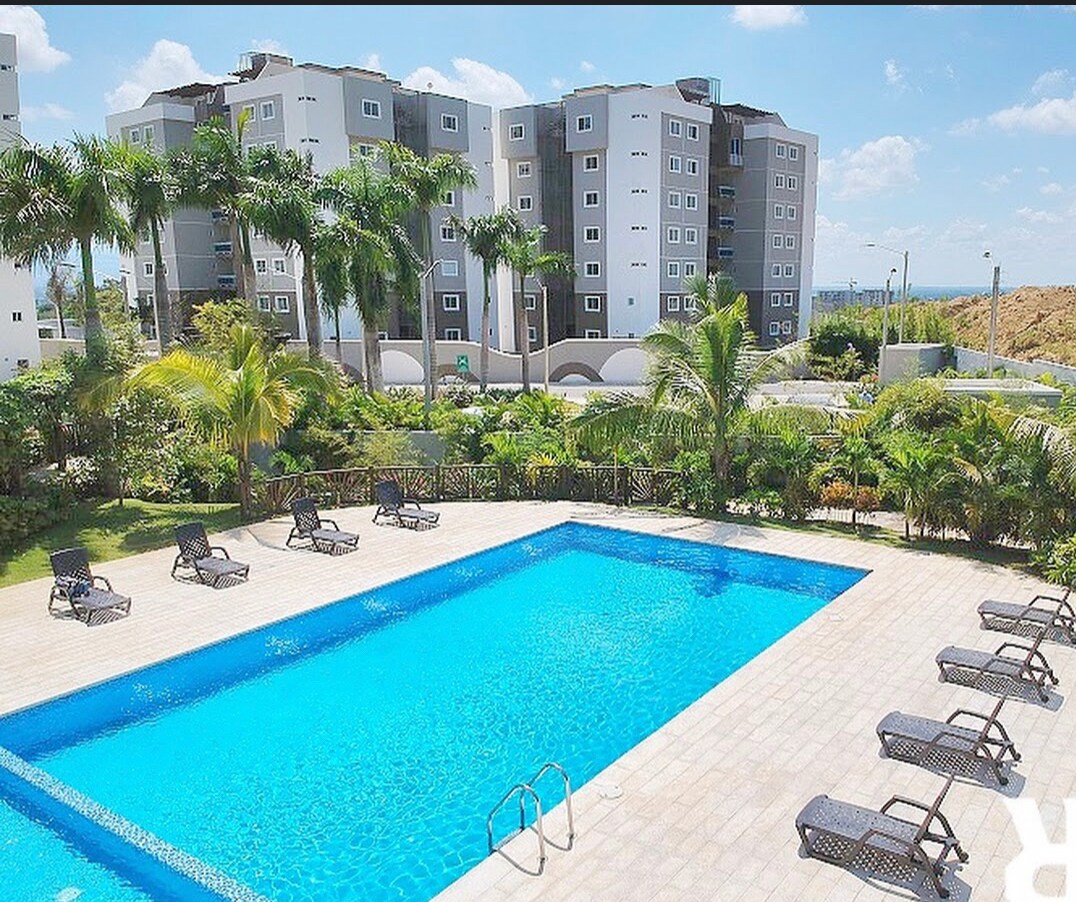 A clear view of a swimming pool is presented, surrounded by a spacious patio. Several lounge chairs are positioned along the edges of the pool, with palm trees providing a touch of greenery. In the background, modern apartment buildings are visible against a bright sky.