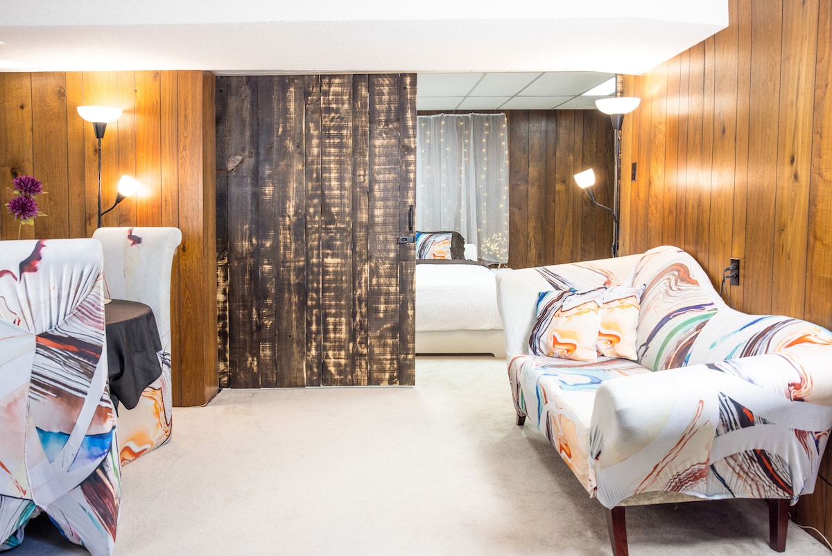 A spacious living area features light-colored wall-to-wall carpet and wooden paneling that adds warmth. Colorful patterned couch covers enhance the seating, while a round dining table sits nearby. Soft lighting is provided by floor lamps, creating a bright and inviting space.