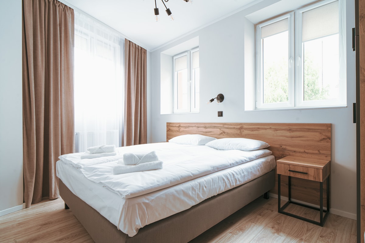 A bright sleeping area features a spacious bed with crisp white linens. Natural light filters through two large windows, complemented by soft, beige curtains. A small bedside table is positioned beside the bed, enhancing the room's functional layout.