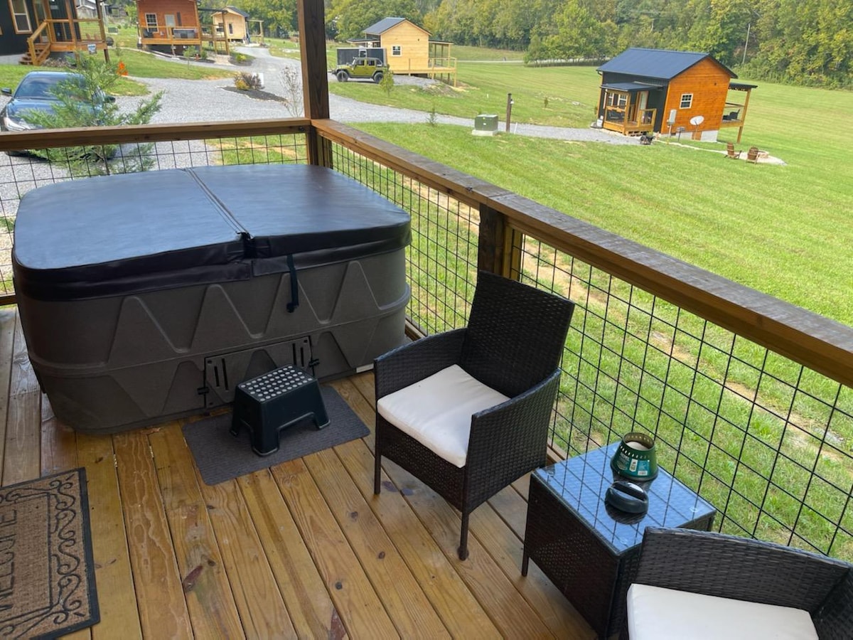 A private deck area is shown with a hot tub, two black wicker chairs, and a small table. A footstool is placed in front of one chair. The surrounding landscape features grassy fields and wooded areas, creating a serene atmosphere.