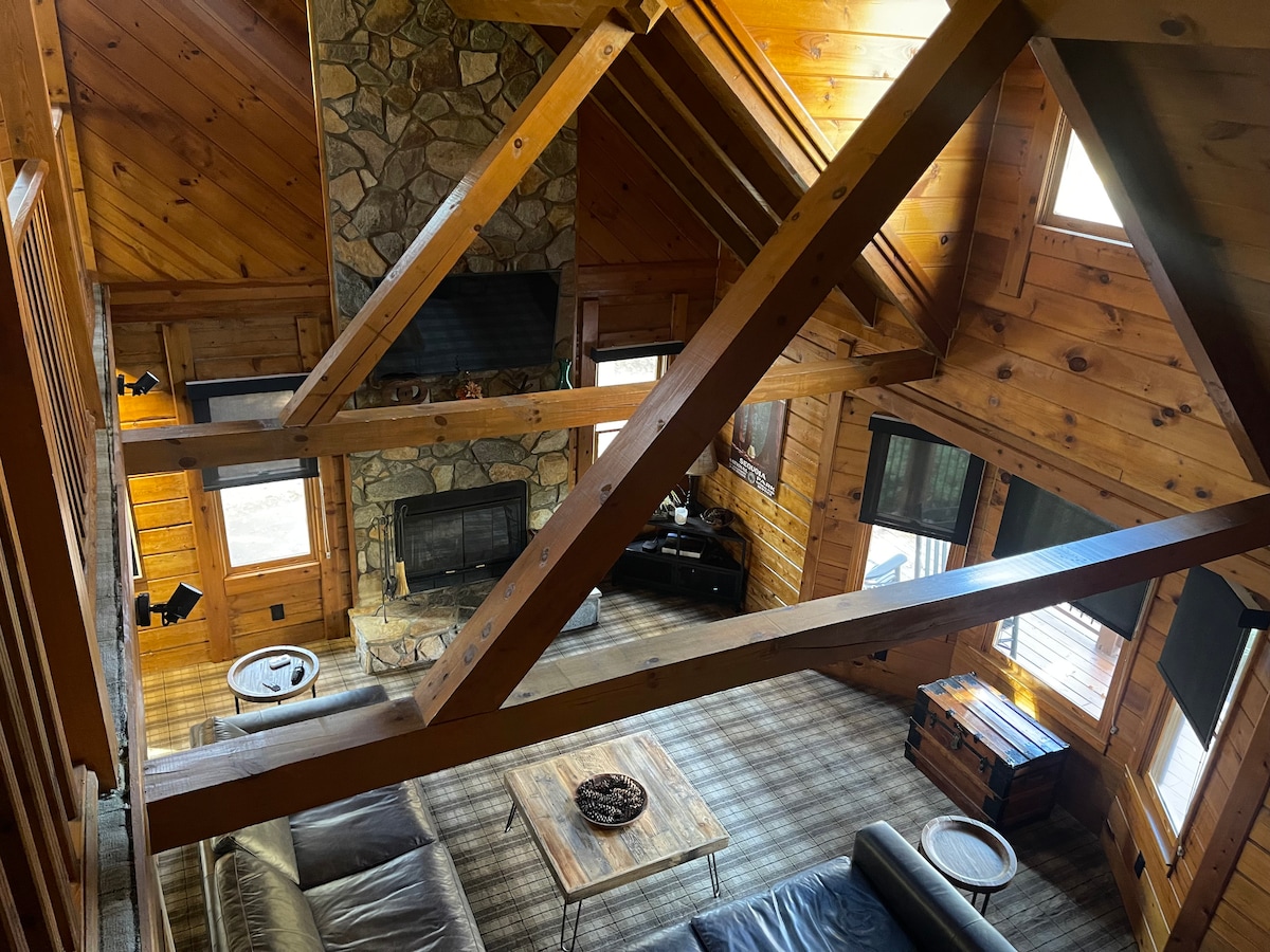 An open living area features a stone fireplace as the focal point, surrounded by large windows that allow natural light to illuminate the wooden walls and ceiling. A cozy seating arrangement, including a dark sofa and coffee table, complements the cabin's rustic charm.