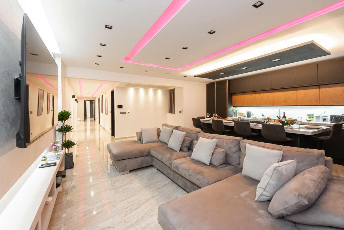 A spacious living area features a U-shaped, luxurious sofa complemented by soft cushions. Mood lighting casts a subtle glow along the ceiling. An open kitchen with modern appliances is visible in the background, enhancing the contemporary design of the space.