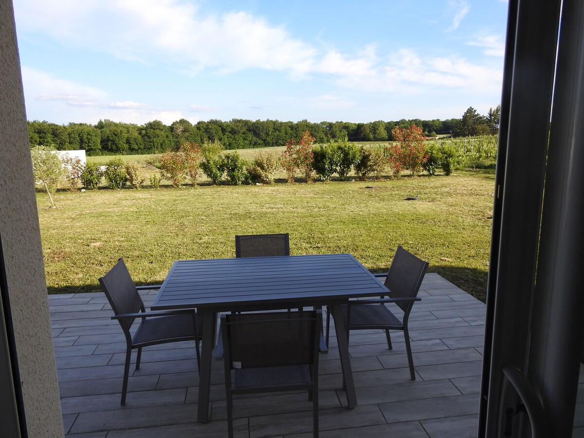 An outdoor patio area is shown, featuring a dining table surrounded by six chairs. A well-maintained grassy lawn extends into a backdrop of trees and shrubs, under a clear blue sky with a few scattered clouds.
