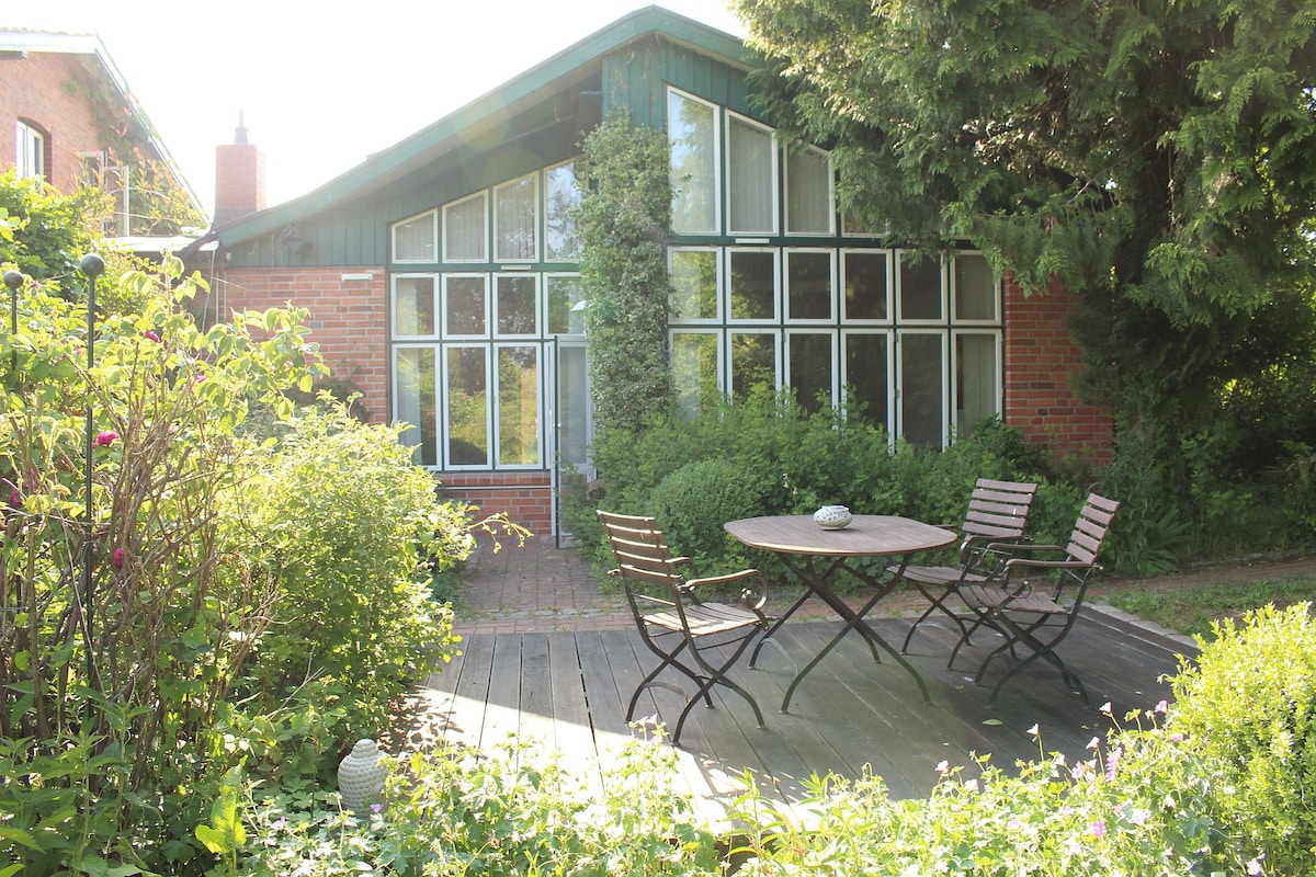 A distinctive house exterior features a large glass front, surrounded by verdant greenery. A wooden deck hosts a round table and four chairs, inviting outdoor relaxation. Sunlight filters through the trees, highlighting the lush garden landscape.