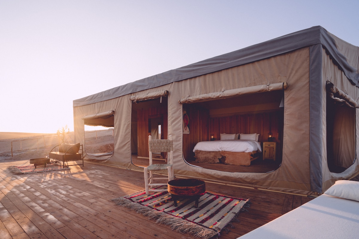 A spacious private tent is set in a serene desert environment, featuring large openings allowing for natural light. A comfortable bed is positioned centrally, complemented by a small round table and a traditional woven rug. The surrounding deck presents a wooden floor with seating options.