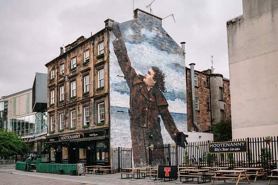 An exterior view of a historic building features a large mural depicting a figure with outstretched arms against a cloudy sky. The mural is prominently displayed on the side of the building, with outdoor seating and a sign for 'Hootenanny' visible in the foreground.