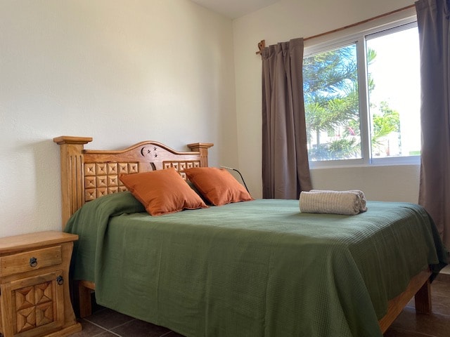 A spacious bed with an earthy green coverlet is positioned against a light wall. Two orange pillows are placed neatly at the head. Natural light enters through a window adorned with beige curtains, revealing a view of trees outside.
