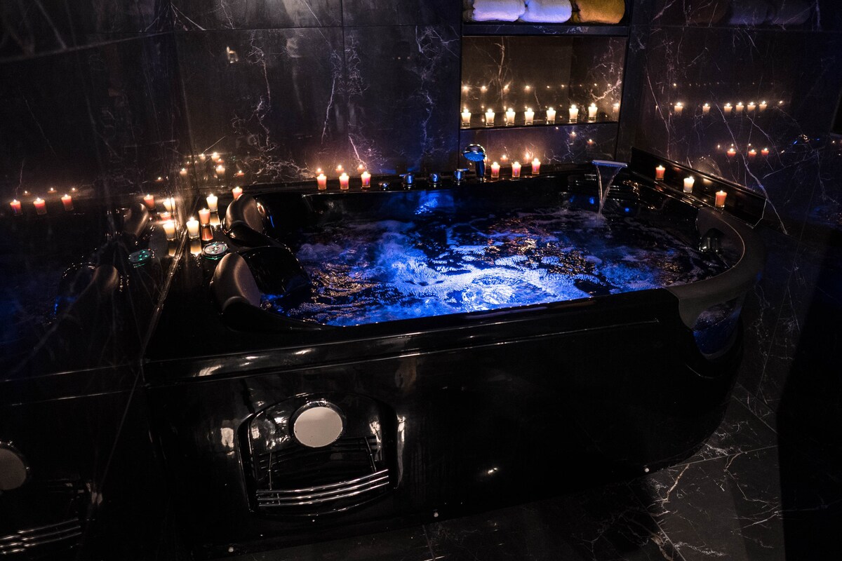 A luxurious jacuzzi is set against a backdrop of dark marble tiles, enhanced by soft lighting from multiple candles. The water reflects a soothing blue hue, creating a calming ambiance within the space.