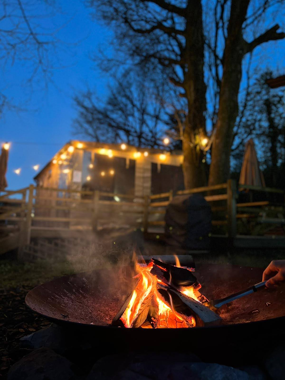 A fire bowl crackles with flames, providing warmth in the evening. Soft lighting from string lights above illuminates the cabin in the background, surrounded by trees and outdoor seating. The scene reflects a cozy outdoor atmosphere, perfect for relaxed gatherings.