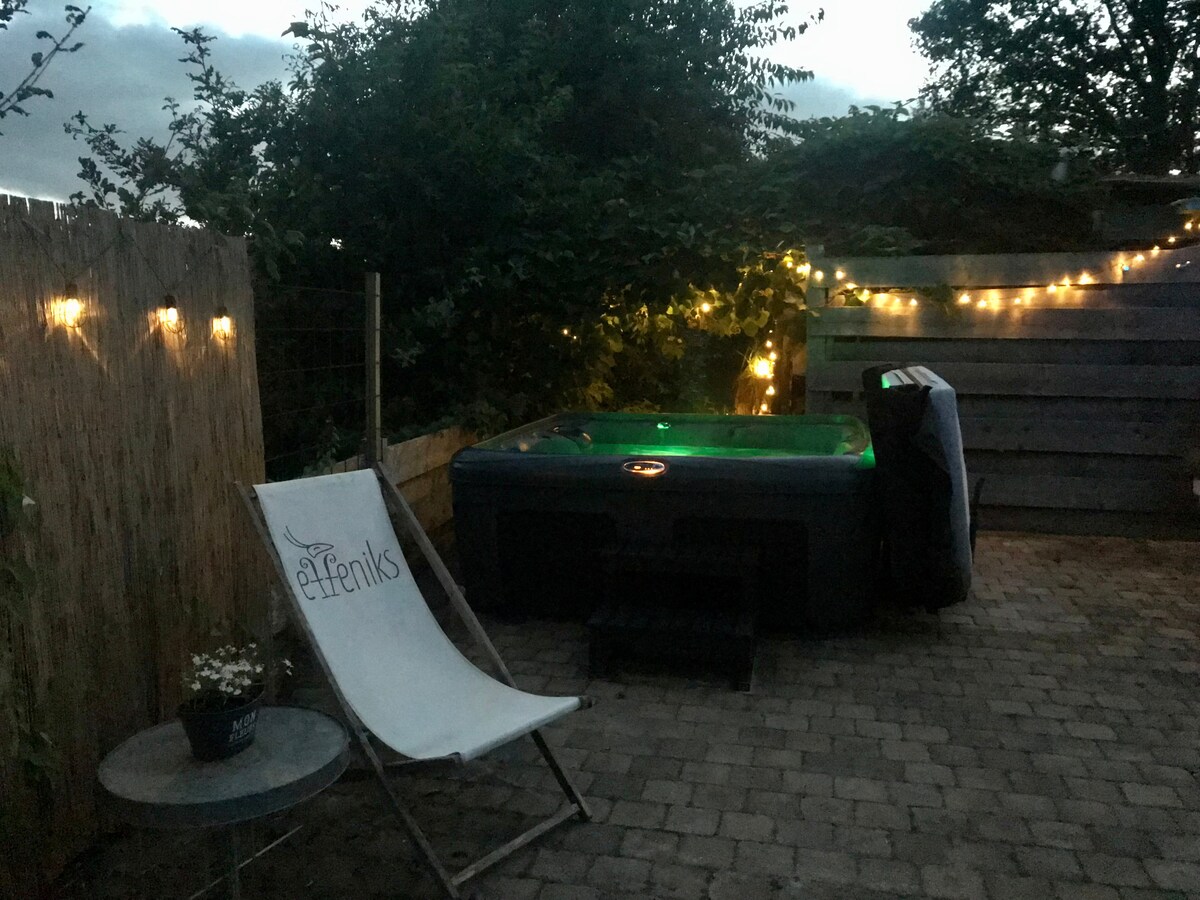 A private outdoor area features a hot tub surrounded by greenery and soft lighting along the fence. A comfortable lounge chair is positioned nearby on a stone patio, creating an inviting space to relax in the evening.