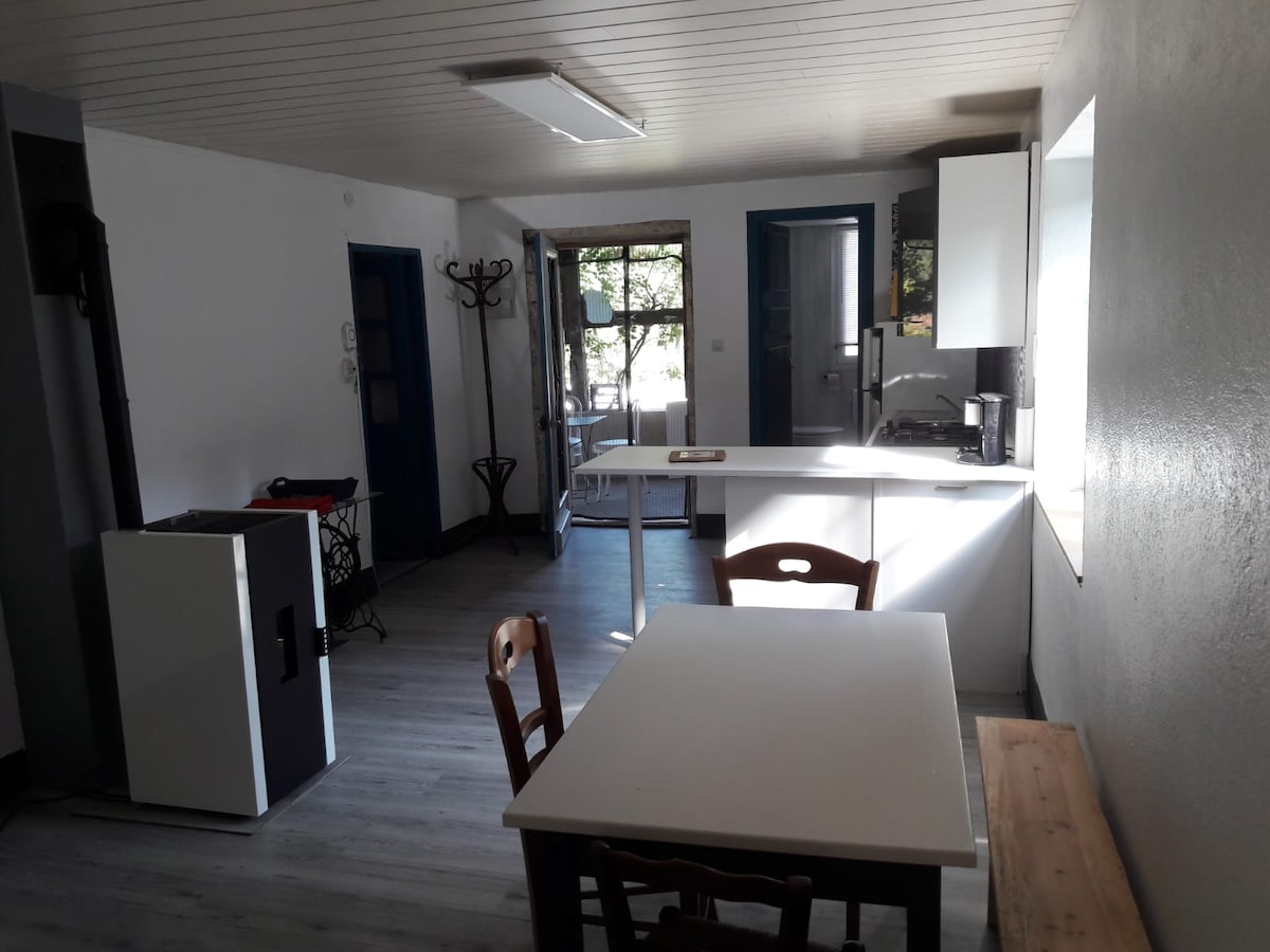 A modern kitchen area is presented with a minimalistic white cabinetry and a spacious countertop. A dining table with wooden chairs is positioned nearby. Natural light enters through a large window, enhancing the openness of the space. A fireplace is visible in the corner.