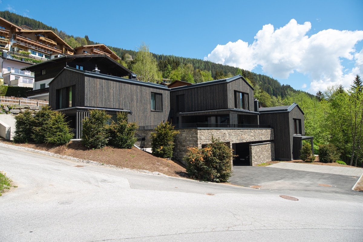 A modern chalet structure is showcased with a combination of wooden and stone exteriors. Surrounded by greenery, the building features large windows and a driveway leading to an integrated garage. The sloping hillside and distant mountains contribute to the serene setting.