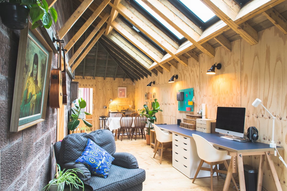 A spacious open-plan area is showcased with wooden beams overhead and large skylights. A cozy seating arrangement is present, along with a work desk featuring a computer. A dining table is set for several guests, complemented by potted plants for a fresh ambiance.