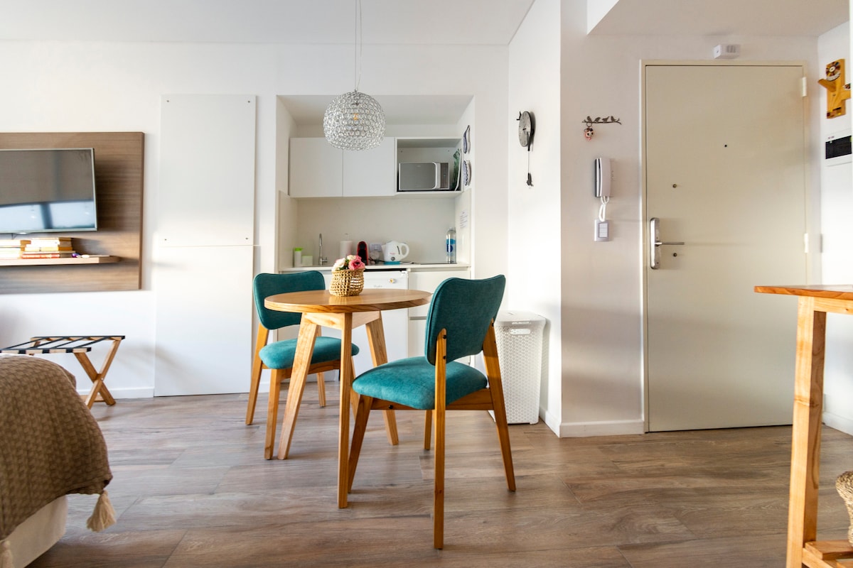 A modern living space is displayed with a round wooden table surrounded by three teal chairs. A compact kitchenette is visible in the background, featuring essential appliances. A smart TV is mounted on the wall, and a glass light fixture hangs from the ceiling.
