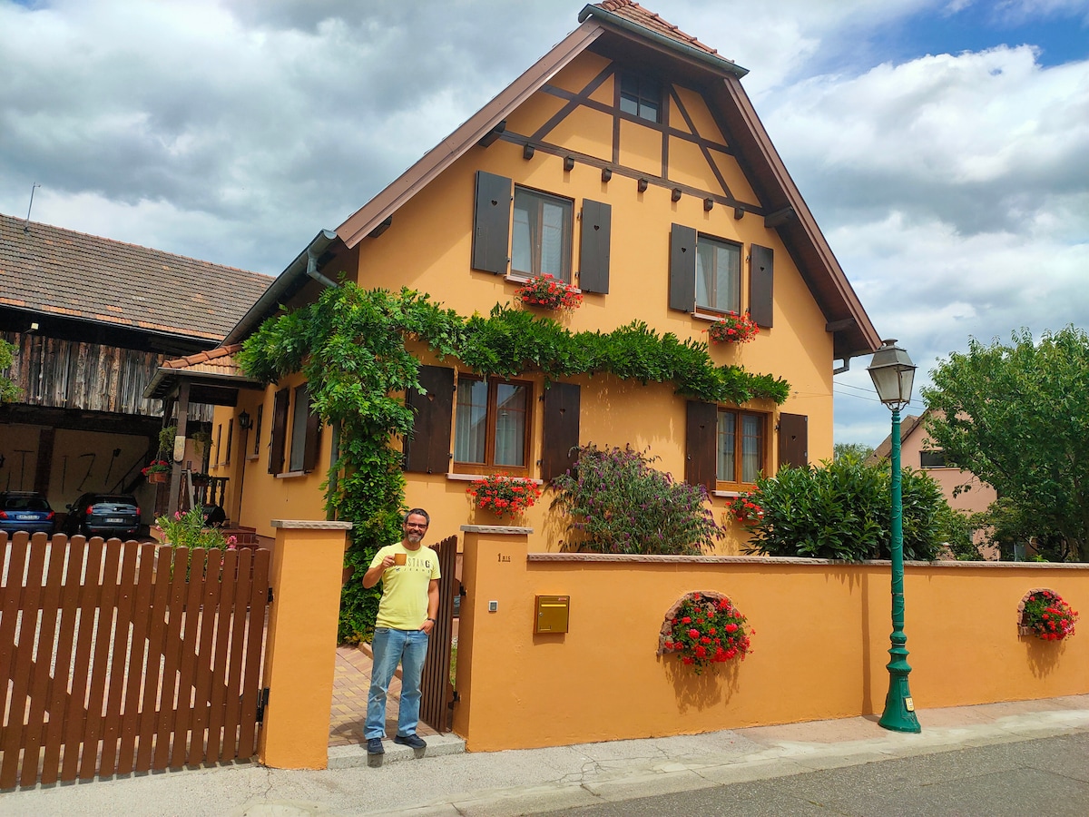 A charming yellow house features decorative wooden shutters and flower boxes filled with vibrant blooms. Lush greenery adorns the facade, enhancing its welcoming appearance. The property is surrounded by a low wooden fence and a cobblestone pathway leading to the entrance.