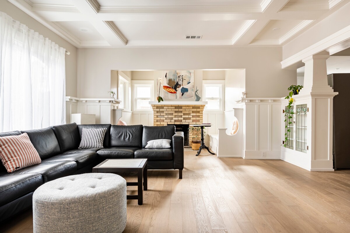 A large living area features a sectional sofa in dark upholstery, accompanied by a round ottoman. A coffee table sits centrally, while an artwork is displayed above a brick fireplace. Windows allow natural light to brighten the space, complemented by white curtains.