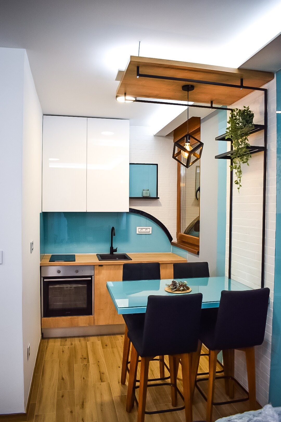 A modern kitchen area features sleek cabinetry and a contrasting wooden countertop. A dining table with four high-backed chairs is positioned nearby. The space is complemented by decorative elements, including a small plant hanging from the ceiling and stylish lighting fixtures.