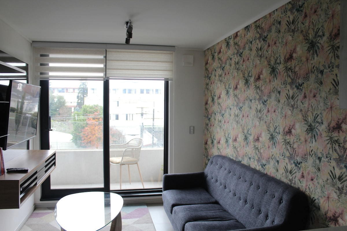 A modern living area features a comfortable sofa and a round coffee table, complemented by a floral accent wall. Large glass doors open to a small balcony, allowing natural light to brighten the space. A TV is mounted for entertainment.