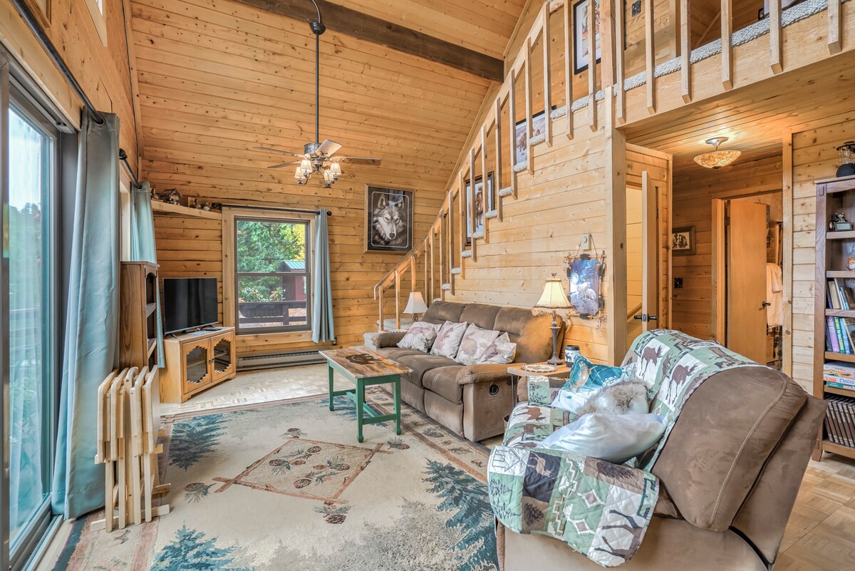 A warm and inviting living area features wooden walls and a high ceiling. Comfortable seating is arranged around a central coffee table, with light filtering through large windows. A staircase leads to a loft space above, while a television sits on a stand nearby.
