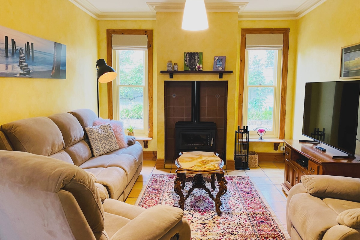 A cozy living room is presented with two soft sofas positioned around a wooden table. A warm-colored wall complements a small fireplace, while natural light filters through two windows. A television sits adjacent, and decorative elements create a welcoming space.