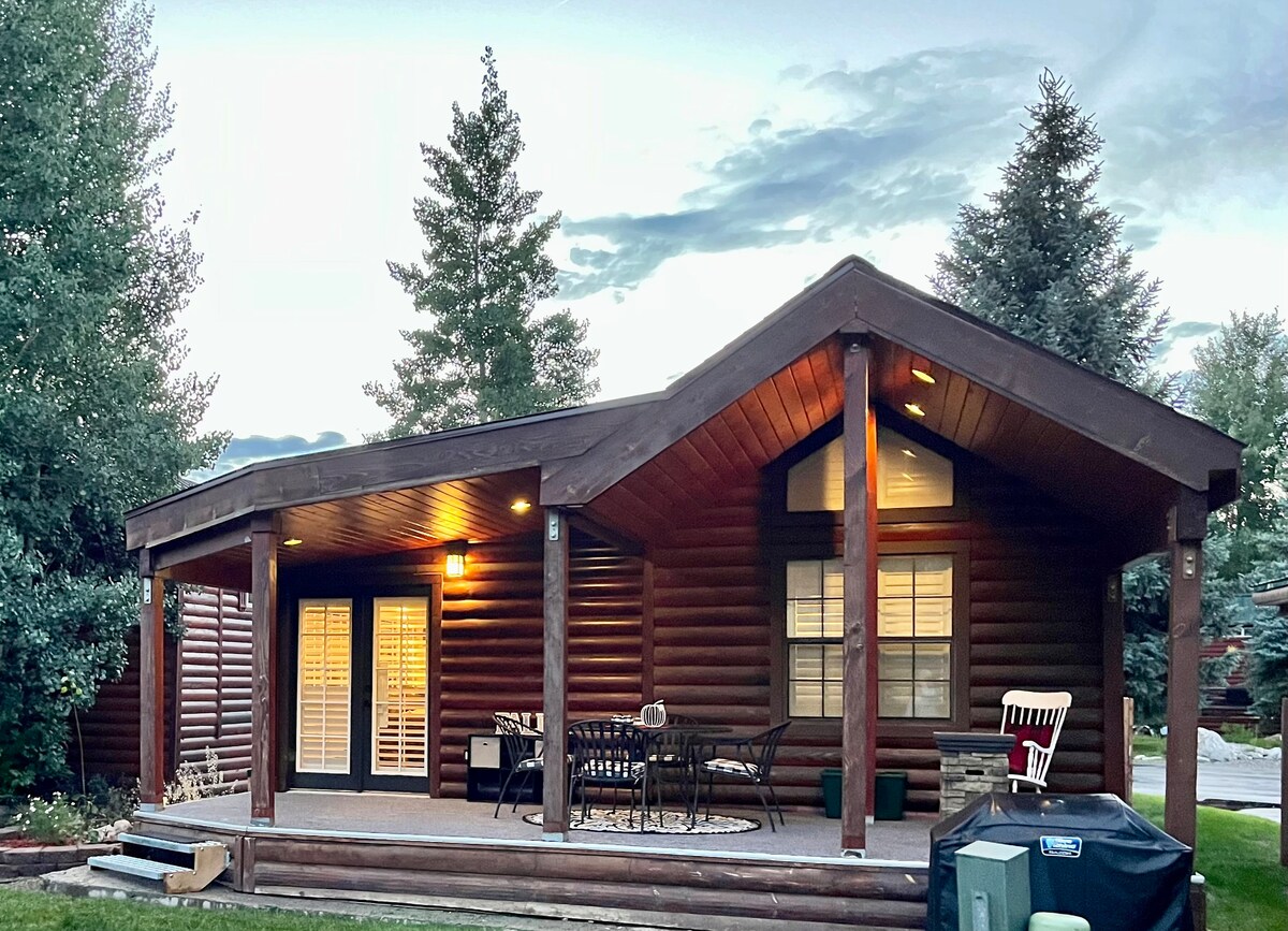 A charming log cabin features a large covered patio with a seating area, surrounded by trees. Soft lighting enhances the inviting atmosphere. The distinct roofline adds character, and large windows allow natural light to brighten the interior.