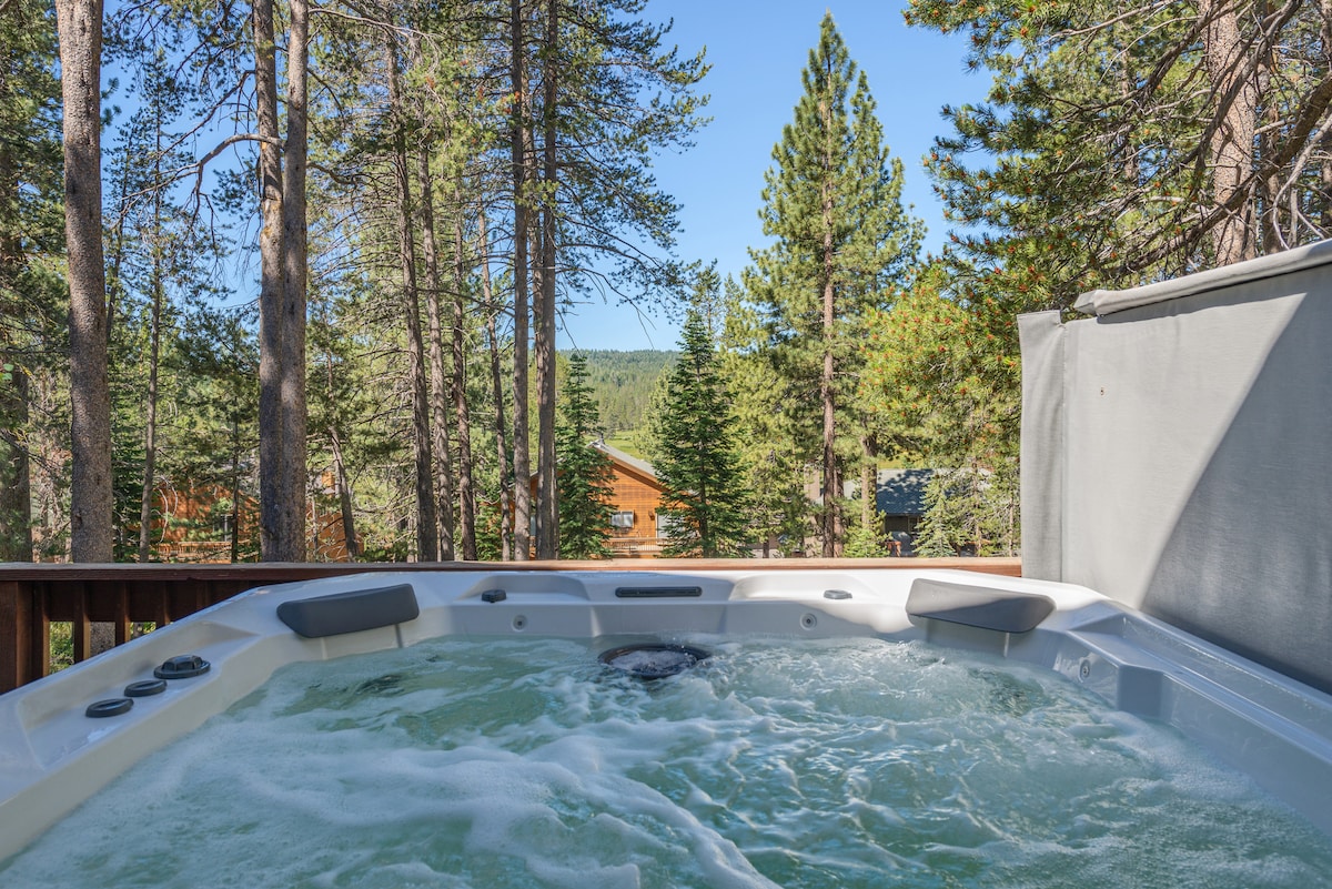 A hot tub is positioned on a wooden deck, surrounded by tall trees and a view of the mountains in the background. Bubbles rise from the water, reflecting the natural light under a clear blue sky.