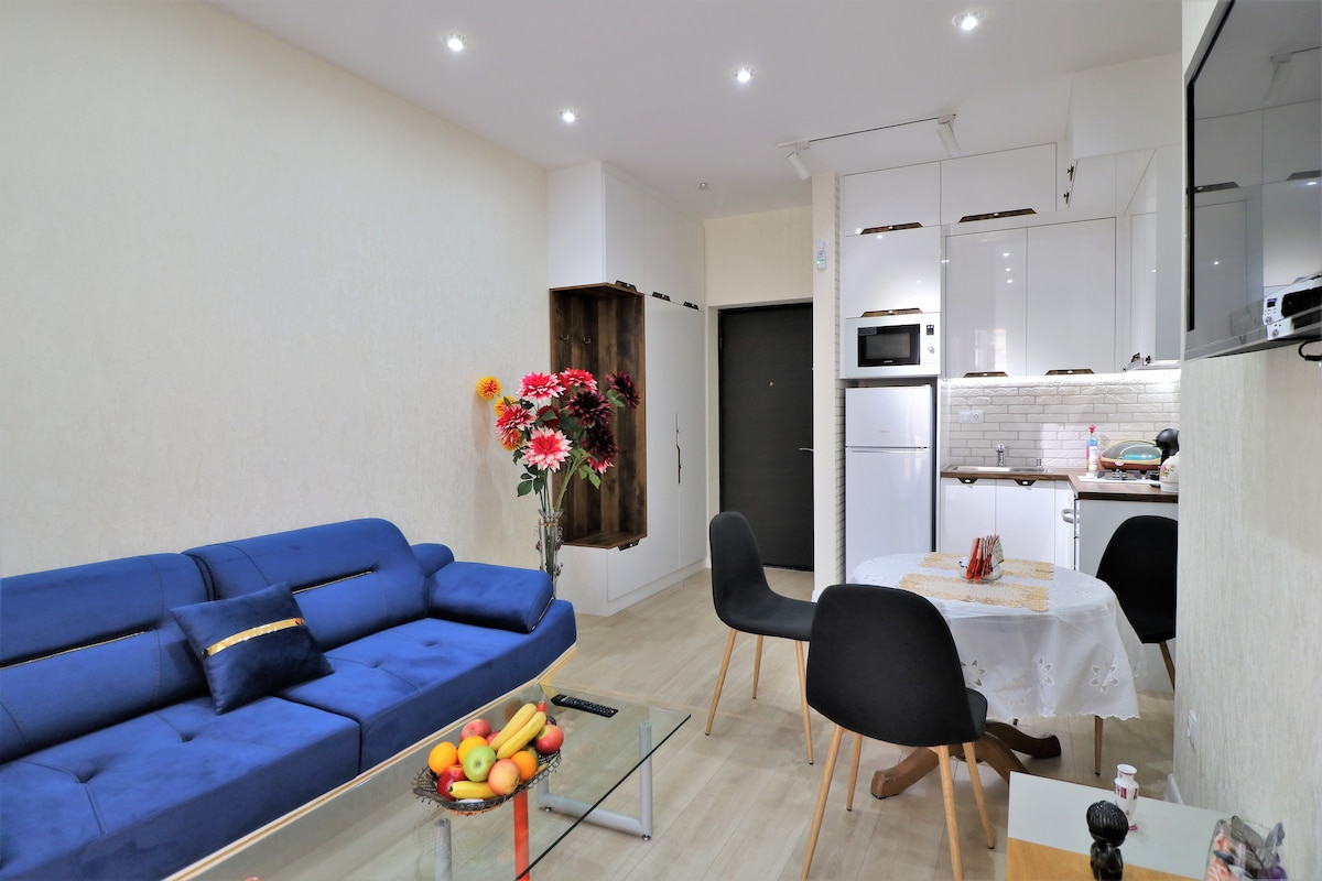 A modern living area features a blue sofa and a glass coffee table adorned with a fruit bowl. A dining table with a lace tablecloth is positioned nearby. The kitchen is designed with sleek white cabinets and includes a microwave and refrigerator, while decorative flowers add a pop of color.