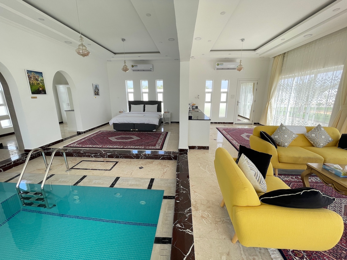 An open-concept space features a private indoor swimming pool adjacent to a spacious seating area. The area is enhanced by large windows allowing natural light, while a bright yellow couch complements the neutral tones of the decor. Generous ceiling heights contribute to an airy atmosphere.