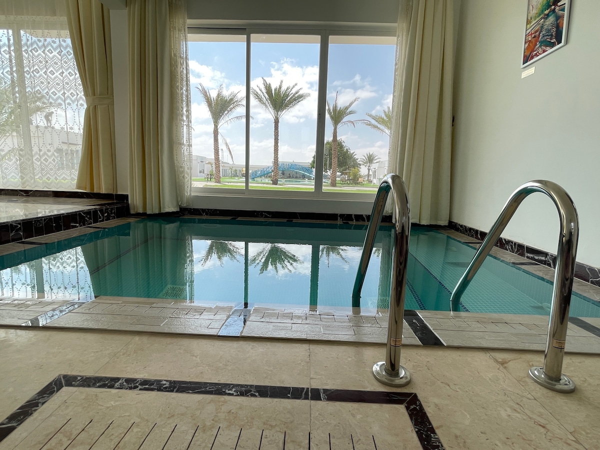 An indoor swimming pool reflects the natural light from large windows, showcasing palm trees and garden views beyond. Metal handrails provide access to the pool, which is surrounded by elegant tiled flooring. Soft drapes frame the windows, enhancing the serene atmosphere.