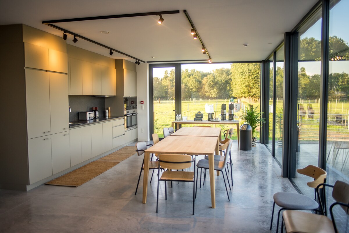 A spacious kitchen and dining area features modern cabinetry and stainless steel appliances. A large wooden dining table is surrounded by a mix of chairs, while large windows provide views of the surrounding fields and outdoor seating. Natural light fills the open space.