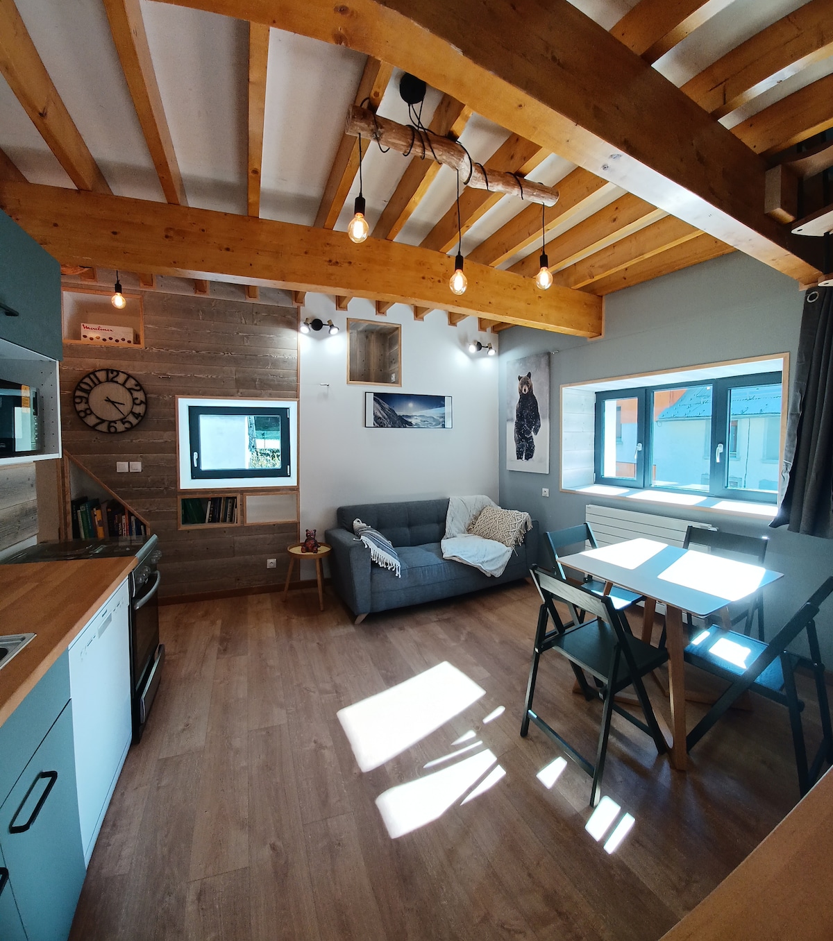 A bright living area features exposed wooden beams and large windows allowing natural light. A cozy sofa is positioned next to a dining table and chairs. A clock and artwork are displayed on the walls, adding character to the inviting space.
