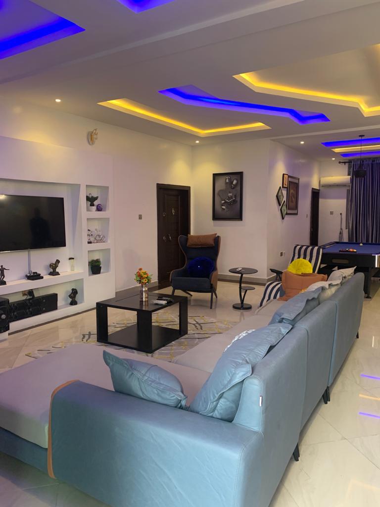 A spacious living area features modern furnishings in neutral tones. A large sectional sofa is complemented by accent chairs and a sleek coffee table. Decorative wall art and a mounted television are visible, with ambient lighting enhancing the room's design.