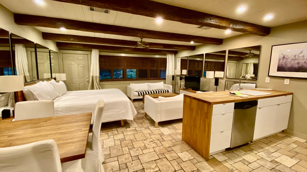 A spacious studio features a King size bed with a white quilt, positioned near a cozy seating area that includes a white sofa and striped accent chair. A modern kitchen island with a wooden countertop and stainless steel appliances is visible. Natural light enters through large mirrors and windows, highlighting rustic wooden beams overhead.