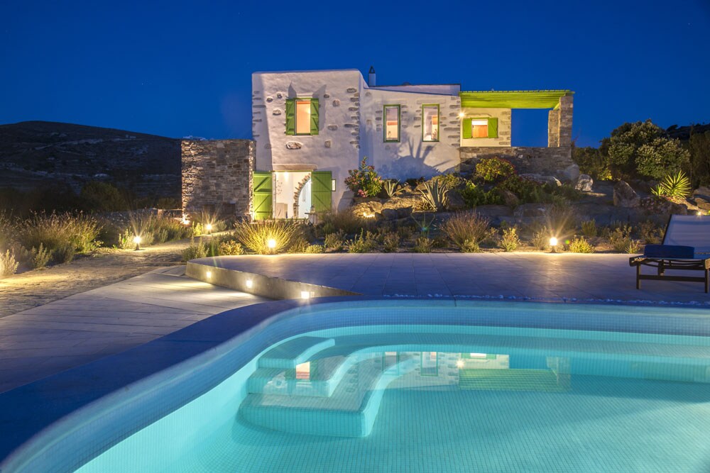 The villa is illuminated at night, highlighting its traditional stone design and green accents. The outdoor area features a swimming pool reflecting the lights, surrounded by landscaped gardens. A serene atmosphere is created by the soft glow of lighting around the premises.
