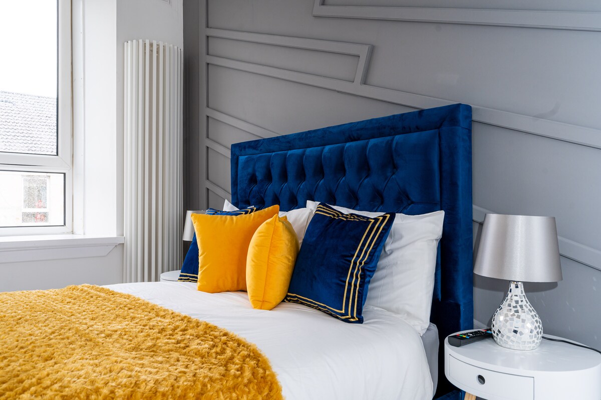 A master bedroom features a luxury upholstered double bed dressed with hotel quality linen. Bright accent pillows enhance the bedding's aesthetic. A stylish nightstand and lamp complement the modern decor, while natural light flows in through the window, highlighting the geometric wall design.