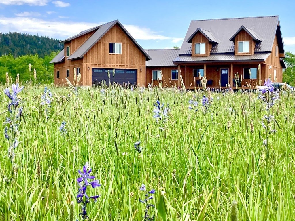 A spacious vacation cabin is set amidst lush green grass, with vibrant wildflowers in the foreground. The cabin's wooden exterior features a large garage and multiple gabled roofs, blending harmoniously with the scenic landscape of hills in the background.