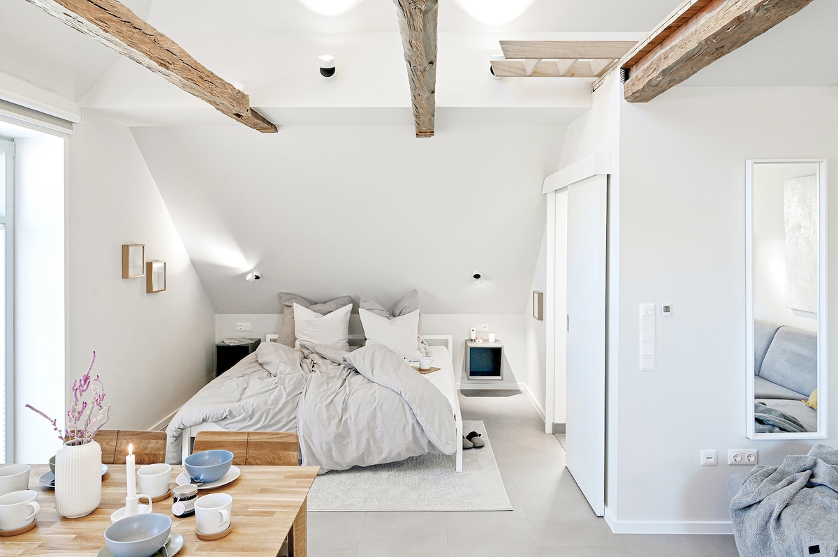 The loft-style interior showcases a spacious sleeping area featuring a large bed with soft linens. Natural light floods the room through two windows, while exposed wooden beams add character. A dining table is set nearby, and a minimalist decor enhances the serene atmosphere.