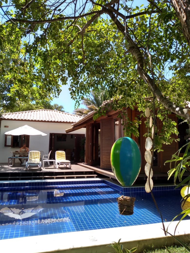 A charming outdoor area features a swimming pool with deep blue tiles, surrounded by a wooden deck. Comfortable lounge chairs offer seating under a large umbrella. A vibrant surfboard is hung from a tree, adding a playful touch to the lush, green surroundings.