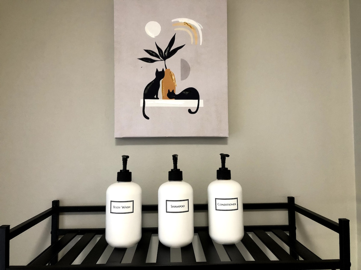 A wall-mounted artwork features an abstract design with two cats and a plant, set above a black storage shelf. Three large white bottles labeled "Body Wash," "Shampoo," and "Conditioner" are displayed symmetrically on the shelf, providing essential toiletries.
