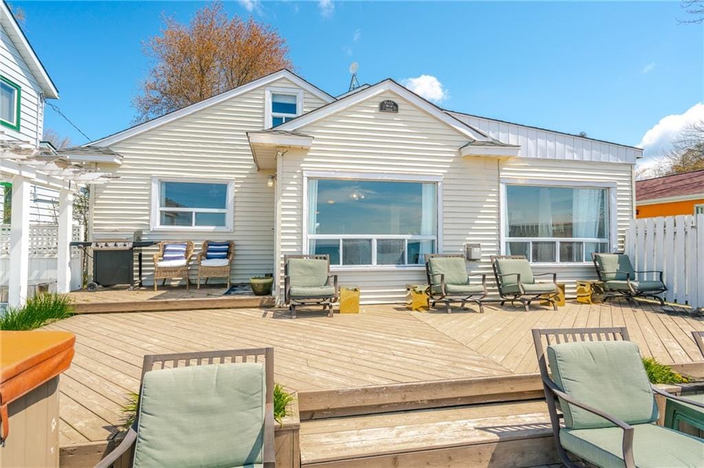 A bright exterior view of the home reveals large windows and a spacious wooden deck adorned with reclining chairs. A grill is positioned nearby, while a clean white fence surrounds the property, creating a welcoming outdoor area for relaxation.