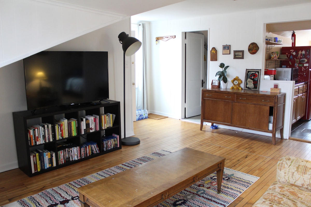 A spacious living area features a large television mounted above a low bookshelf filled with books and movies. A wooden coffee table sits at the center, complemented by a light-colored couch. Doorways lead to other areas, with a hint of the kitchen visible in the background.