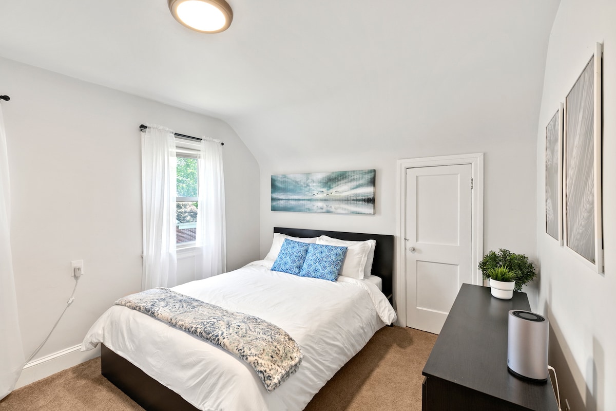 A bright bedroom features a spacious bed dressed in white linens and a decorative blanket. Natural light enters through sheer curtains, complementing the neutral wall color. A dresser and a small plant add subtle touches, while artwork hangs above the bed, contributing to a calm ambiance.