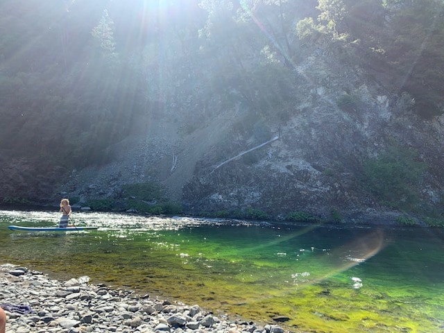 A serene river scene is depicted with a person quietly paddling on a stand-up paddleboard. Sunlight streams through the trees, creating a gentle shimmer on the water's surface, which transitions from clear to vibrant green near the shoreline lined with smooth stones.