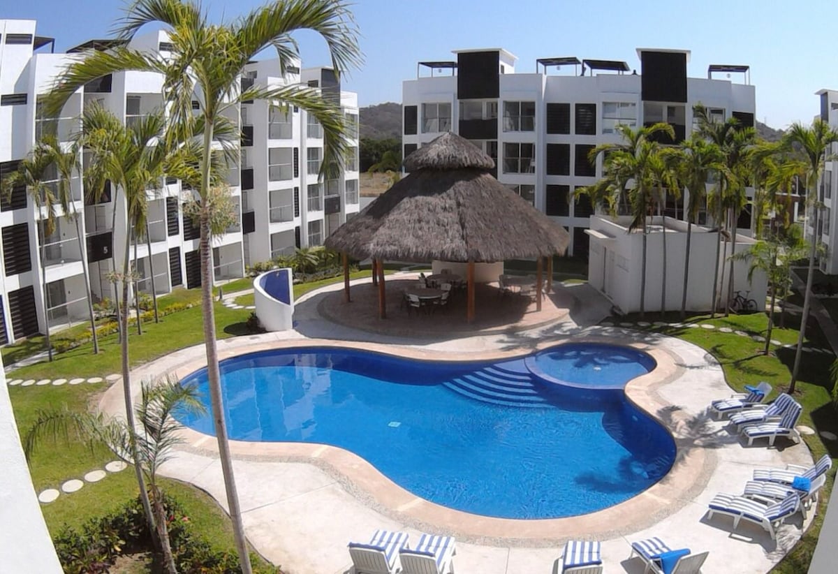 A sunlit pool area is surrounded by well-maintained green lawns and palm trees. A thatched-roof palapa offers shaded seating nearby, while several lounge chairs are arranged around the pool. The modern condominium buildings frame the space, creating a relaxing atmosphere.