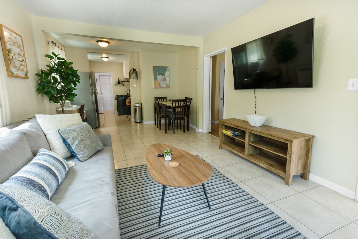 A spacious living area features a gray sofa adorned with blue and white cushions, paired with a circular wooden coffee table atop a striped area rug. A mounted television is visible on the wall, and a dining table is located in the background, with greenery enhancing the space.